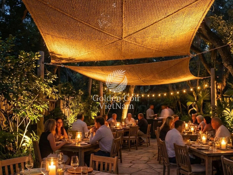 Golden-brown coconut coir shade sails with warm uplighting over an outdoor restaurant, creating a rustic luxury evening dining ambiance.