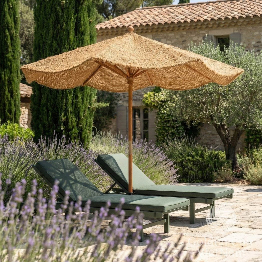 Two green lounge chairs under a large Coco umbrella in a garden setting with lavender and a stone building.